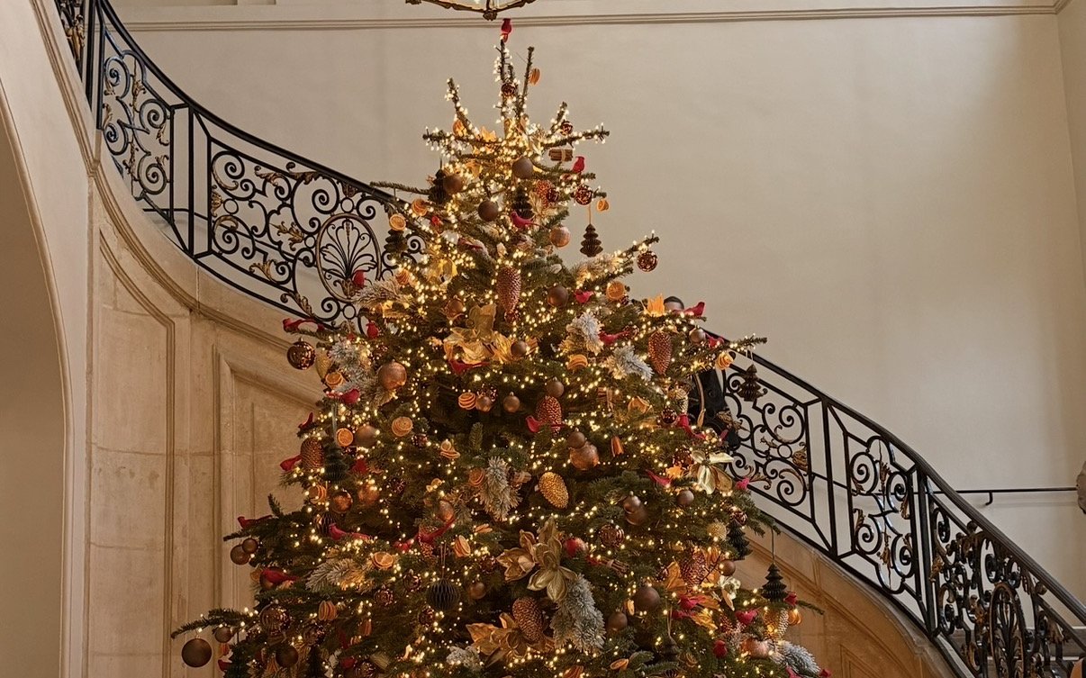 A tall Christmas tree in gold and copper lights beneath a lantern, at the foot of a sweeping wrought-iron staircase